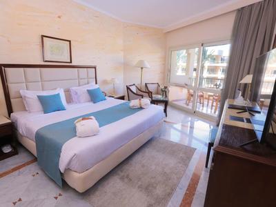 Bright hotel room with large bed, TV, chairs, and balcony door letting in natural light.