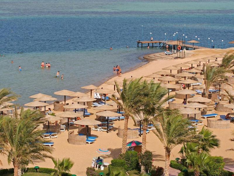 Sunny beach with palm trees, umbrellas, and people relaxing near the calm sea.