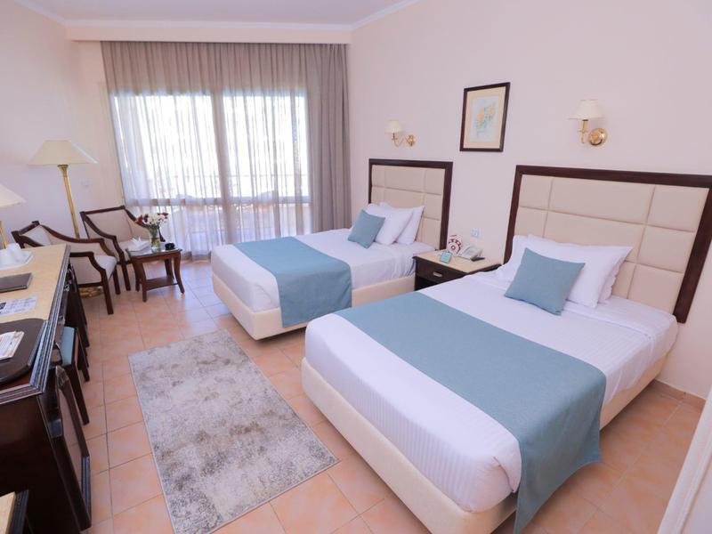 Hotel room with two single beds, light blue accents, chairs, desk, and large window.