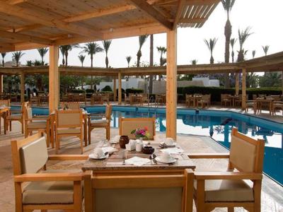 Venkovní restaurace s dřevěnými židlemi a výhledem na bazén pod dřevěnou střechou.