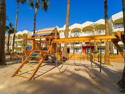 Holz-Kinderspielplatz mit Schaukeln und Klettergerüst in sonnigem, palmengesäumtem Hof.