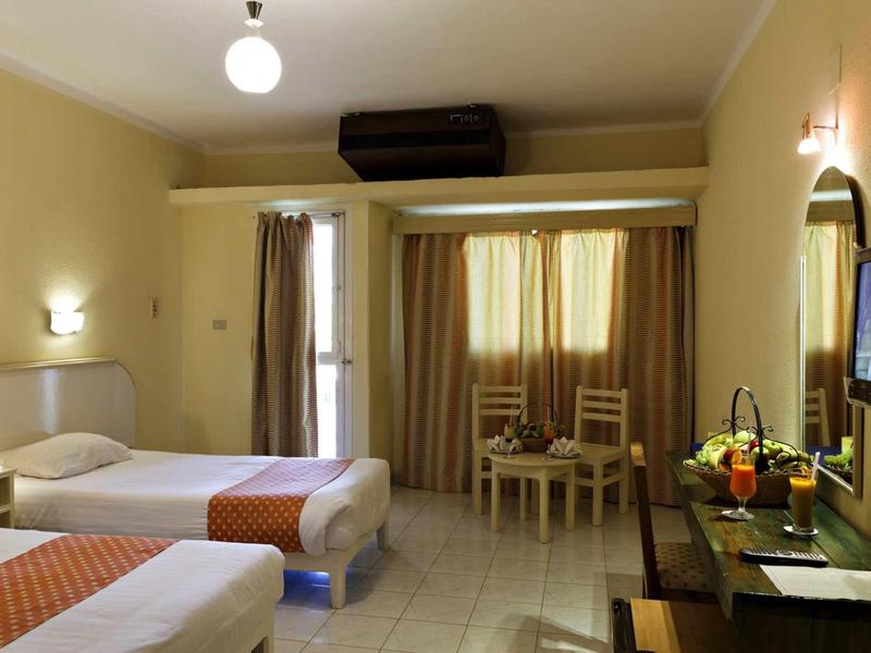 Jasny pokój hotelowy z dwoma łóżkami, stołem z krzesłami, telewizorem i zasłonami na oknie.