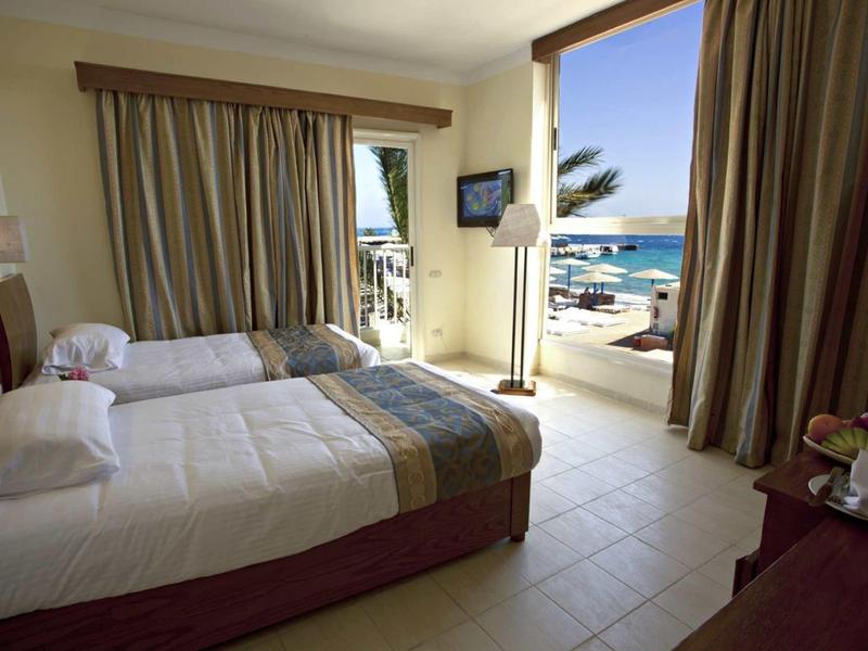 Pokój hotelowy z dwoma łóżkami, zasłonami, balkonem i słonecznym widokiem na morze.