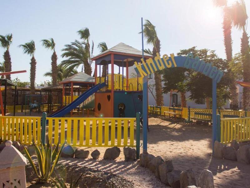 Kinderspielplatz mit gelbem Zaun, Klettergerüst, Palmen und Sandboden bei Sonnenlicht.