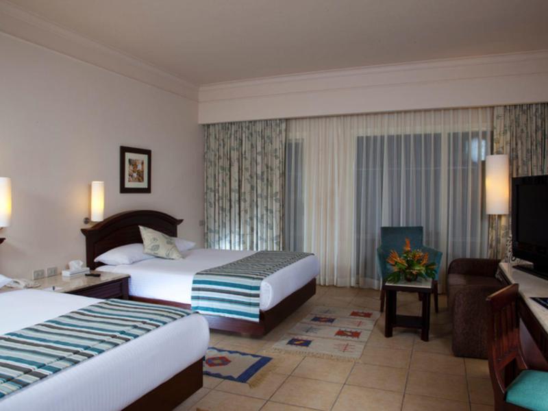 Hotelový pokoj se dvěma jednolůžky, nočními stolky, křesly a balkonem se závěsy.