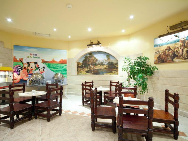 Světý interiér s dřevěnými židlemi a stoly, nástěnnými malbami a rostlinami v hotelové části.