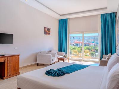 Bright hotel room with double bed, armchairs, and balcony overlooking green grounds.