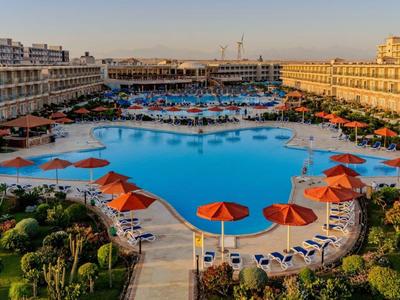 Large hotel pool with red umbrellas, surrounded by multi-story buildings at sunset.