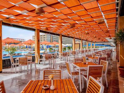 Covered terrace with wooden tables and chairs overlooking a pool and hotel building.