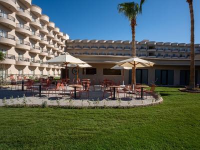 Hotelterrasse mit runden Tischen, Sonnenschirmen und Grasfläche im Vordergrund.