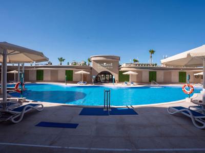 Runder Hotelpool mit Liegestühlen und Sonnenschirmen unter klarem blauem Himmel.