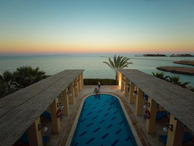 Blick auf langes Schwimmbecken mit Palmen und Meer im Hintergrund bei Sonnenuntergang.