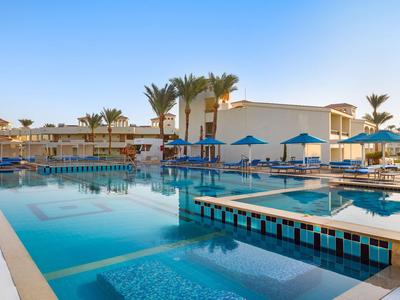Modern outdoor pool area with blue umbrellas and palm trees in front of a hotel building under clear sky.