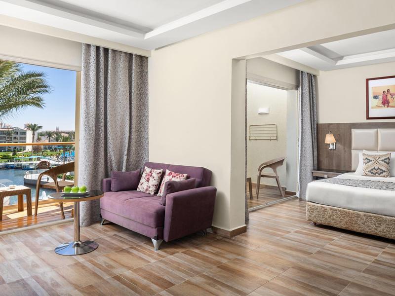 Nowoczesny pokój hotelowy z sypialnią, częścią wypoczynkową i widokiem na basen i palmy.