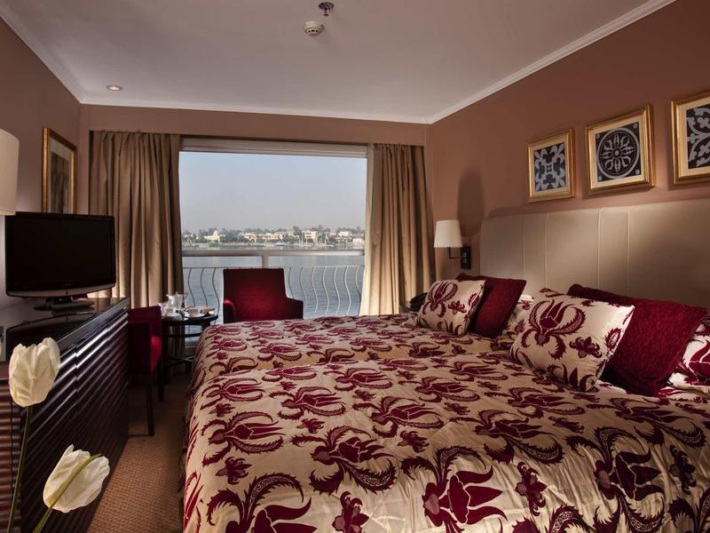 Przytulny pokój hotelowy z dużym łóżkiem, ozdobnymi poduszkami, telewizorem i widokiem na port.