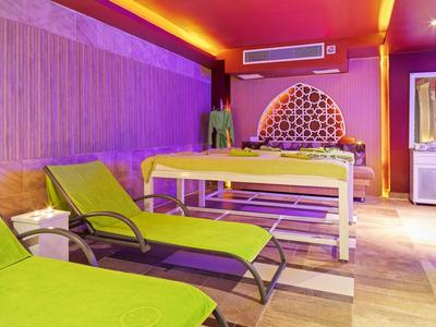 Salone spa con lettini da massaggio e sedie verdi sotto luce viola e arancione.