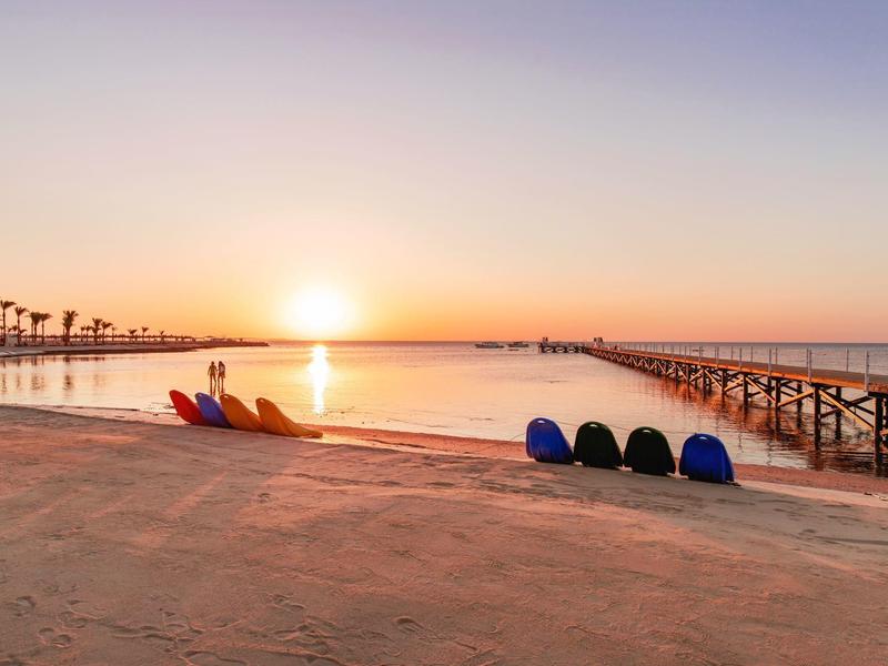 Strand bei Sonnenaufgang mit bunten Kajaks und langem Steg im ruhigen Wasser.