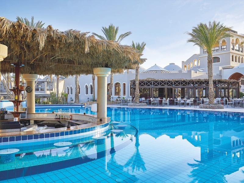 Grande piscine avec palmiers et parasols en paille dans un complexe hôtelier ensoleillé.