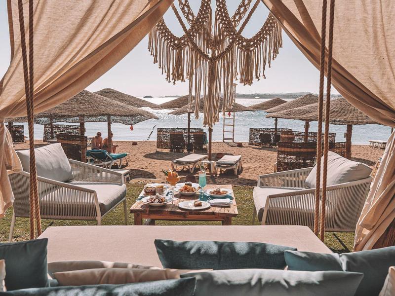 Lounge sulla spiaggia di lusso con cuscini, tende e lampada sospesa decorativa con vista sul mare.