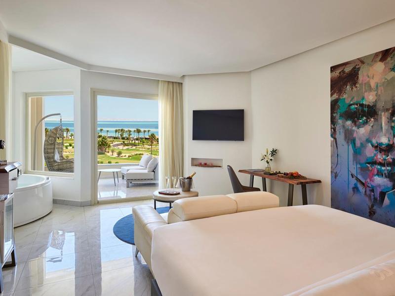 Habitación de hotel luminosa con cama grande, balcón con vista al mar y un cuadro moderno en la pared.