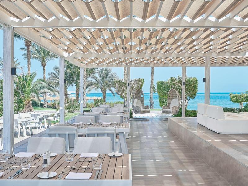 Área de comedor al aire libre moderna y abierta con vista al mar y palmeras bajo un techo de persianas de madera.
