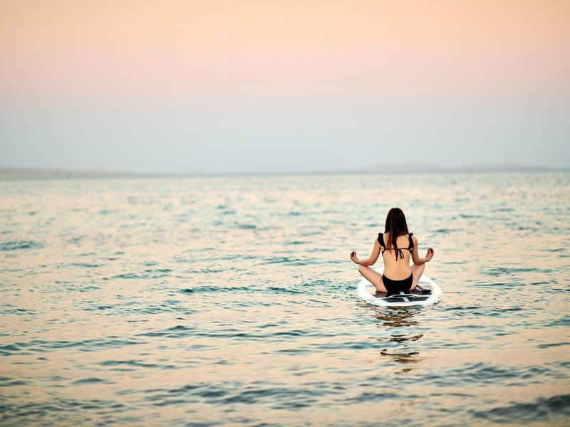 Persona sentada meditando en una tabla de paddle en el mar tranquilo al atardecer.