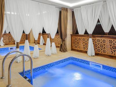 Piscina interna con illuminazione blu circondata da tende e pannelli murali decorativi.