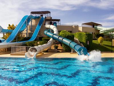 Wasserrutschen führen in einen großen Pool in einer sonnigen Urlaubsanlage.