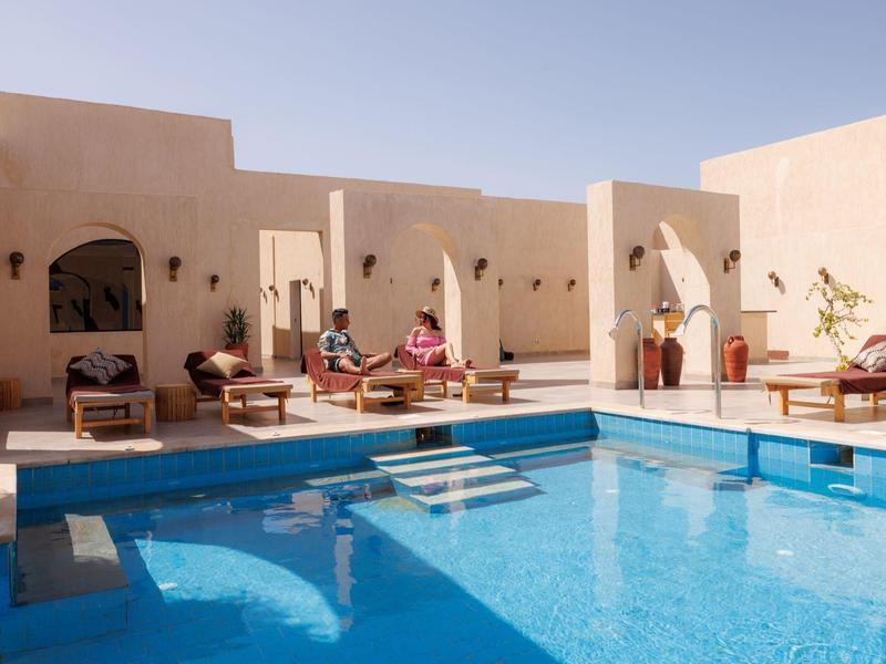 Innenhof mit klarem blauem Pool, Liegestühlen und sandfarbenen Häusern im sonnigen Licht.