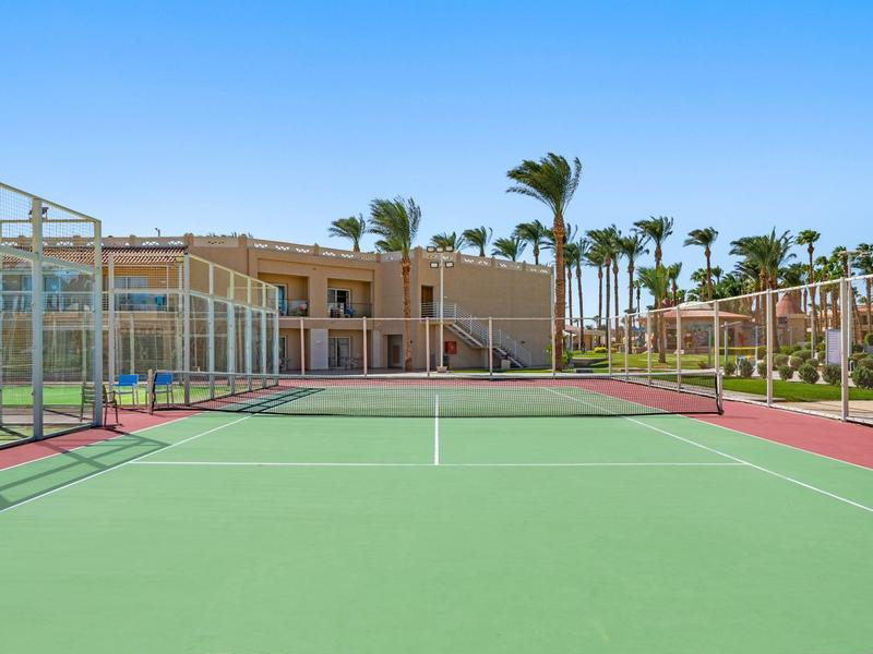 Pista de tenis en un complejo hotelero con edificios y palmeras al fondo bajo un cielo despejado.