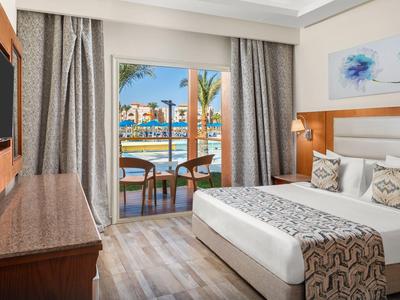 Lichte hotelkamer met tweepersoonsbed, patroon kussens en balkon met uitzicht op palmbomen en zwembad.