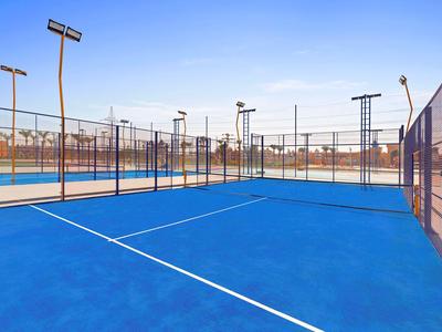 Een leeg blauw tennisveld met hekken en meerdere schijnwerpers onder een heldere lucht.