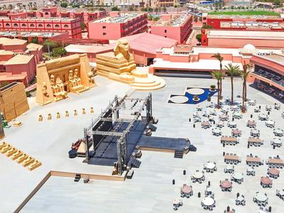 Ampia piazza con statue dorate e tavoli sotto ombrelloni rossi e blu.
