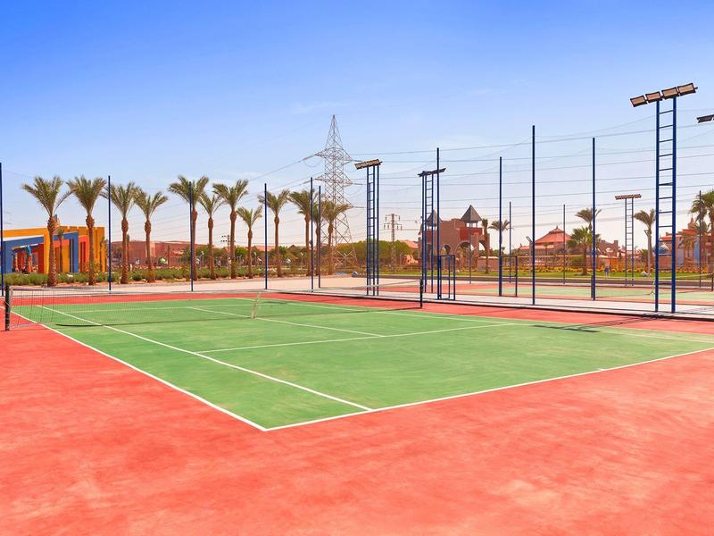 Campo da tennis all'aperto con recinzioni e palme sotto un cielo soleggiato.