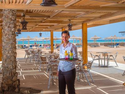 Serveuse servant des boissons dans un restaurant en bord de plage ensoleillé avec sable et parasols à l'arrière.
