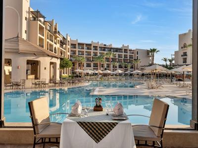 Une table dressée avec vue sur une grande piscine d'hôtel et des parasols sous un ciel dégagé.