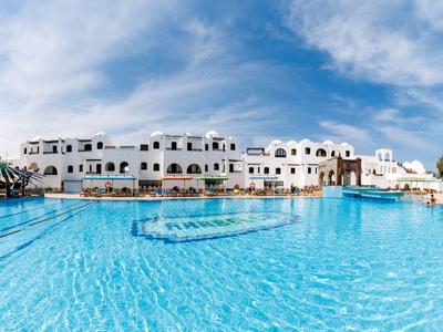 Velký venkovní bazén s modrou vodou před bílou hotelovou budovou pod modrou oblohou.