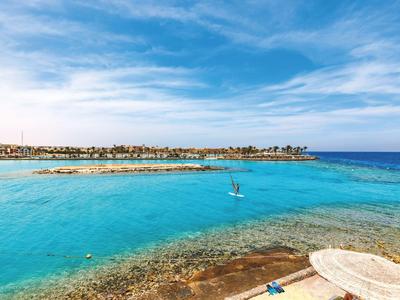 Pohled na křišťálově čistou vodu a pobřeží se dvěma windsurfaři pod modrou oblohou.