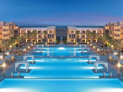 Großer beleuchteter Hotelpool mit modernen Gebäuden und Palmen bei Abenddämmerung.