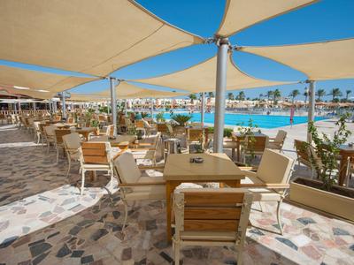 Freiluftrestaurant mit Tischen, Stühlen und Schattensegeln neben einem Pool bei klarem Himmel.