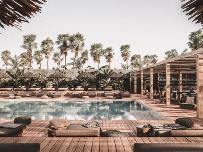 Outdoor hotel pool area with loungers, palm trees, and shaded seating under wooden pergolas.