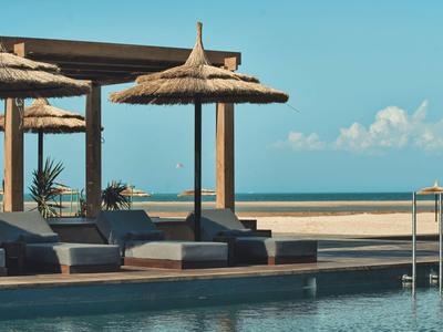 Poolbereich mit Sonnenschirmen, Liegen und Blick auf den Strand und das Meer bei klarem Himmel.