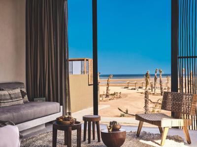 Moderner Wohnbereich mit großem Fenster und Blick auf einen Sandstrand und blauen Himmel.