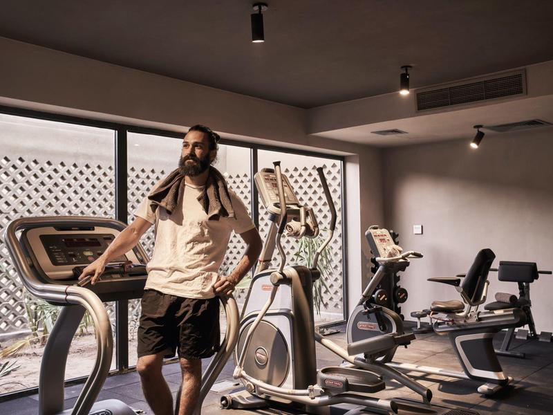 Gym room with exercise equipment lit by large windows and a person on an elliptical trainer.
