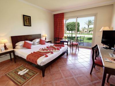 Helles Hotelzimmer mit zwei Betten, Fliesenboden, Schreibtisch, TV und Blick auf Garten mit Terrasse.