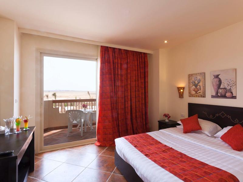 Helles Hotelzimmer mit Doppelbett, roten Vorhängen, Balkon und Blick auf das Meer.