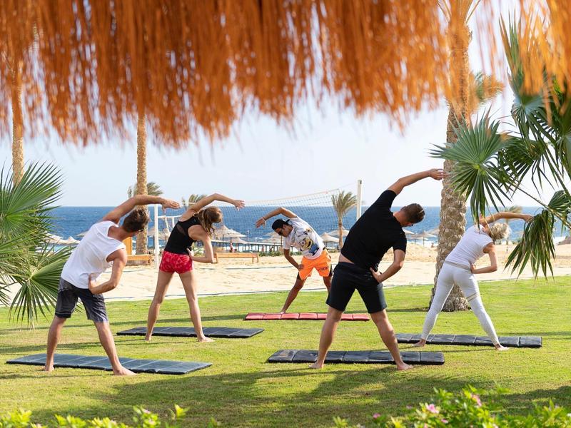 Fünf Personen machen Yoga auf grüner Wiese am Strand mit Palmen und Sonnenschirmen im Hintergrund.