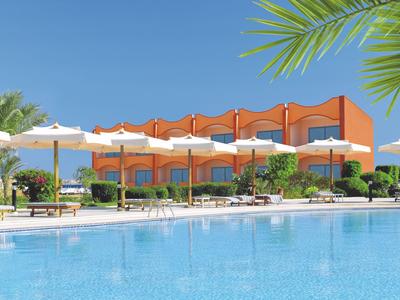 Un hotel moderno con fachada naranja junto a una gran piscina y sombrillas bajo un cielo despejado.