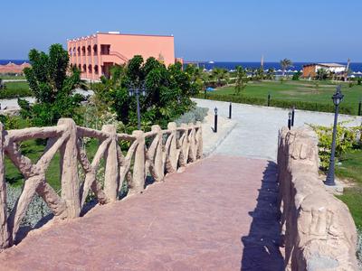 Path with wooden railing leading to a peach-colored building near the coastline on a sunny day.