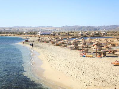 Playa con agua clara, arena y sombrillas de paja junto a un alojamiento turístico.